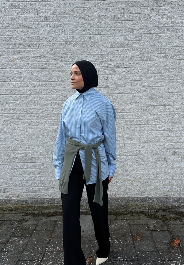 Une femme portant un hijab noir, une chemise à rayures bleues, un pantalon noir et des chaussures blanches se tient près d’un mur de briques gris et regarde sur le côté.