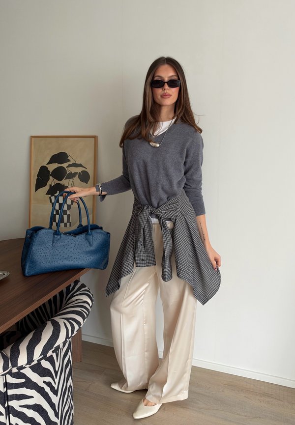 Une femme portant des lunettes de soleil présente un pull gris, un pantalon crème et une chemise à carreaux nouée autour de la taille. Elle tient un sac à main bleu près d’un bureau.