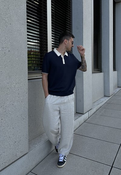 Polo navy con cuello crema, combinado con pantalones de lino claros y zapatillas negras con detalles en blanco. Entorno urbano con ventanas.