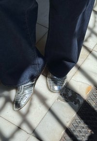 Ezüst fényű, texturált, hüllőszerű mintákkal ellátott cipők, amelyeken két fekete vízszintes csík található, sötét farmer bővülő nadrággal párosítva.