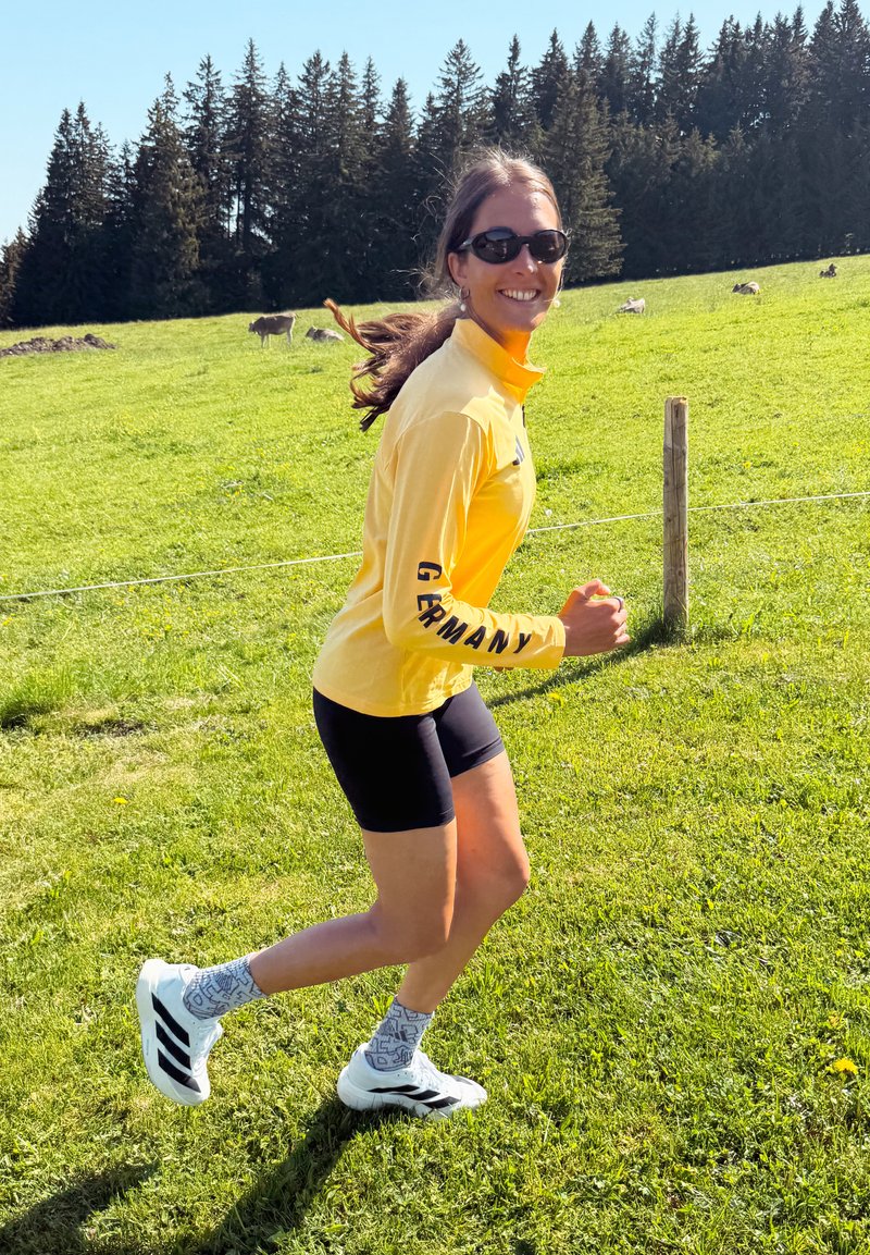 Femme faisant du jogging en extérieur sur un champ d'herbe verte, portant un t-shirt jaune Allemagne, un short noir, des lunettes de soleil, des baskets blanches, avec des arbres et des animaux en pâturage en arrière-plan.