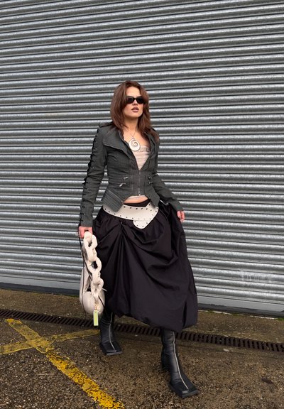 Mujer con chaqueta oscura, falda negra voluminosa, botas negras, gafas de sol, sosteniendo un bolso beige fruncido, de pie junto a una persiana metálica.