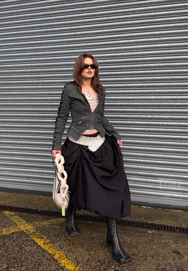 Žena u tamnoj jakni, voluminoznoj crnoj suknji, crnim čizmama i sunčanim naočalama drži bež torbicu s naborima, stojeći uz metalnu roletnu.