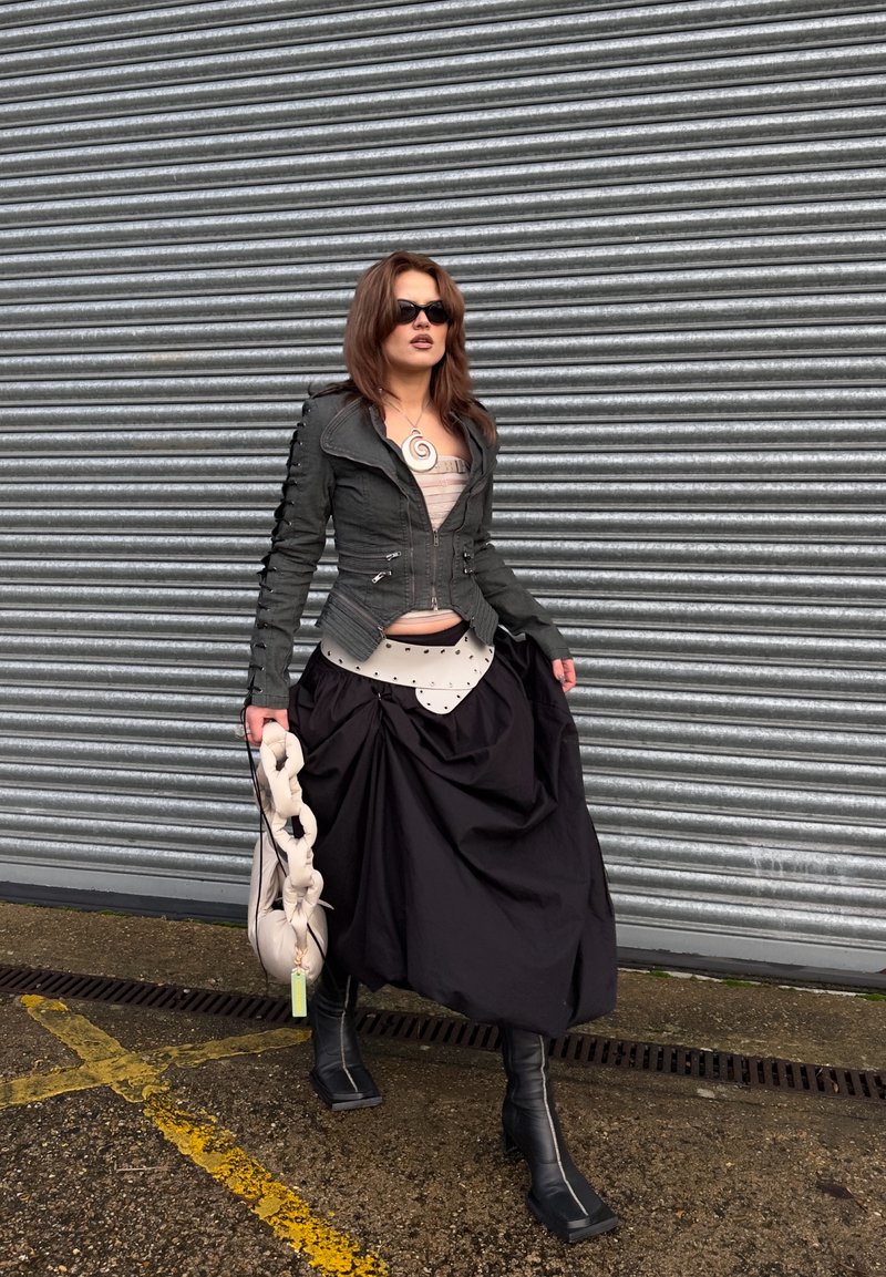 Frau mit dunkler Jacke, voluminösem schwarzem Rock, schwarzen Stiefeln, Sonnenbrille, hält eine geraffte beige Handtasche und steht neben einem Metallrollladen.