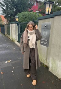 Femme marchant sur un trottoir près d'une porte, portant un long manteau marron, une écharpe beige et marron, un pantalon large et des chaussures beige clair par une journée nuageuse.
