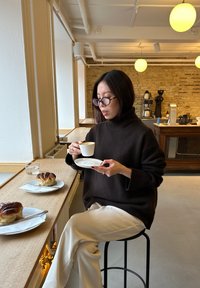 Brun strikket genser med høy hals, hvite bukser, holder en kaffekopp, sitter på en svart krakk ved en trebenk med bakverk.