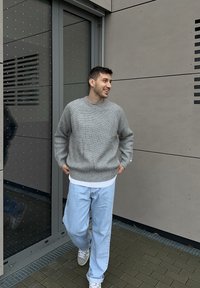 Sivý pletený sveter s textúrovaným vzorom, kombinovaný so svetlomodrými džínsami. Prostredie je zariadené modernými stavebnými materiálmi a dizajnom.