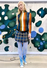 Sieviete ar garām blondām matiem, ģērbusies svītrainā turtleneck, zilā kvadrātveida svārkos, gaiši zilos mežģīņu zeķbiksēs un zilo un baltu sporta apavu komplektā, stāv telpā.