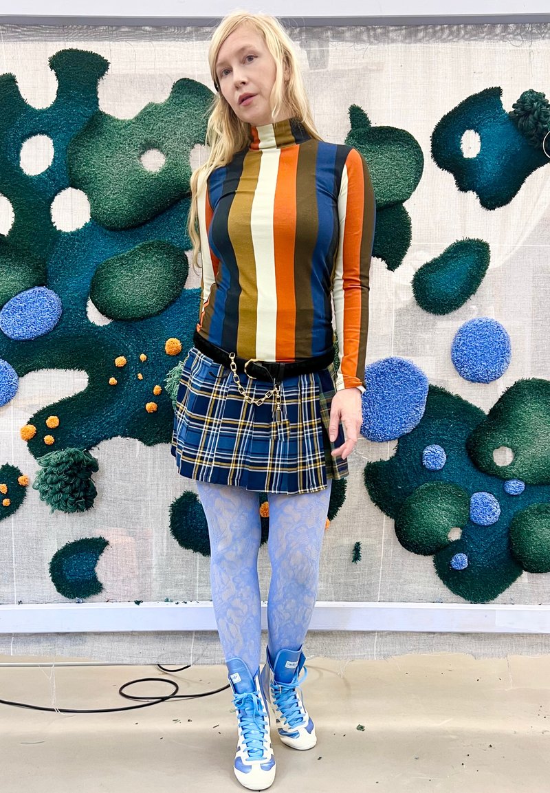 Femme avec de longs cheveux blonds portant un col roulé rayé, une jupe à carreaux bleus, des collants en dentelle bleu clair et des baskets bleu et blanc se tient à l'intérieur.