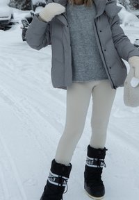 Giubbotto imbottito grigio, maglione grigio chiaro, leggings crema e stivali da neve neri con accenti bianchi. Sfondo innevato. La texture è morbida e calda.