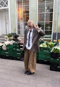 Pārmērīgi liels brūns žakets, balta krekla, melna kaklasaite un platas bēšas bikses. Melni apavi un rokassoma. Stāv priekšā zaļiem dārzeņu kastēm.