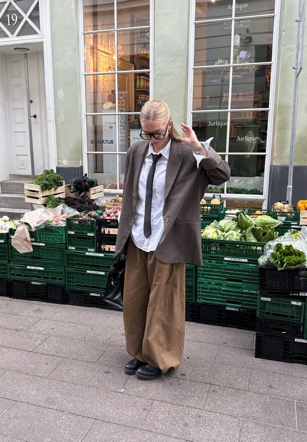 Przesadny brązowy żakiet, biała koszula, czarny krawat i szerokie beżowe spodnie. Czarne buty i torebka. Stoi przed zielonymi skrzyniami na warzywa.