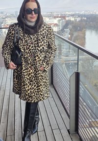 Manteau en fausse fourrure imprimé léopard avec deux boutons, associé à des bottes noires en cuir montant jusqu'aux genoux. La personne tient un sac à main noir et se tient sur un balcon.