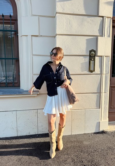 Chaqueta de camisa de mezclilla con botones blancos, combinada con una falda blanca acampanada. Botas altas hasta la rodilla de ante color miel. Bolso de hombro marrón. Fondo claro.