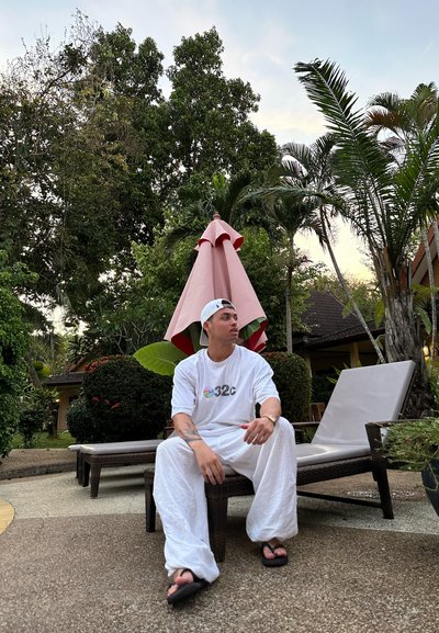 Camiseta blanca oversize con un logo multicolor, pantalones blancos fluidos y chanclas. Un paraguas rosa y plantas tropicales en el fondo.