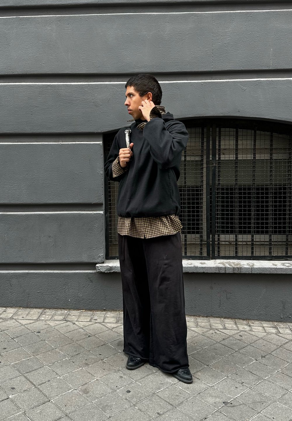 Czarny oversize'owy sweatshirt na brązowej koszuli w kratę, zestawiony z luźnymi czarnymi spodniami i czarnymi butami. Szary mur w tle.