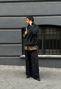 Czarny oversize'owy sweatshirt na brązowej koszuli w kratę, zestawiony z luźnymi czarnymi spodniami i czarnymi butami. Szary mur w tle.