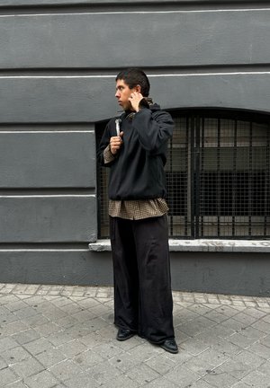Sudadera negra de gran tamaño sobre una camisa de cuadros marrón, combinada con pantalones holgados negros y zapatos negros. Muro gris en el fondo.