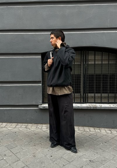 Sudadera negra de gran tamaño sobre una camisa de cuadros marrón, combinada con pantalones holgados negros y zapatos negros. Muro gris en el fondo.