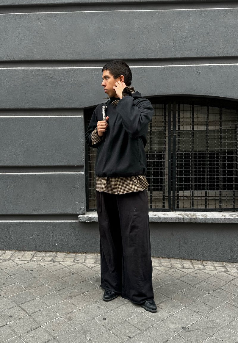 Sweater preto oversized sobre uma camisa xadrez castanha, combinado com calças pretas largas e sapatos pretos. Parede cinza ao fundo.