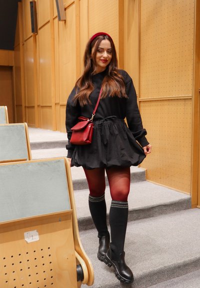 Vestido negro de mangas largas con falda acampanada, combinado con medias marrones y calcetas hasta la rodilla. Accesorios incluyen un bolso rojo y una diadema.