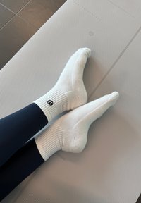 Hvite bomullssokker med ribbestrikkede mansjetter, med en liten svart logo. Føttene er plassert på en teksturert grå matte.