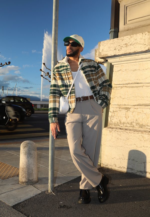 Karierte Flanelljacke in Grün, Beige und Weiß; weißes T-Shirt; beigefarbene Hose; schwarze Plateaustiefel; weiße Umhängetasche; Sonnenbrille; Außenbereich.