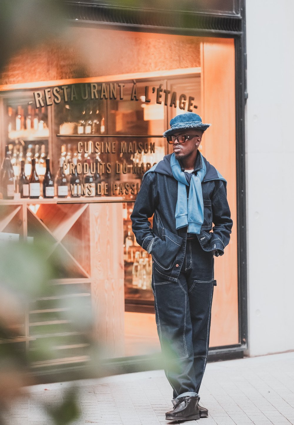 Dark denim jacket and trousers with contrasting stitching, paired with a light blue sweater and a blue hat. Brown shoes complete the outfit.
