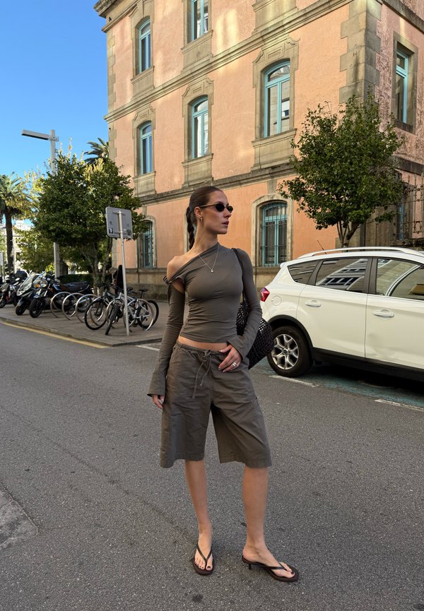 Tumepruun vabaõla off-shoulder pikkade varrukatega pluus, millele on sobitatud laiad khaki lühikesed püksid ja mustad sussid; mudel hoiab musta kootud kotti.
