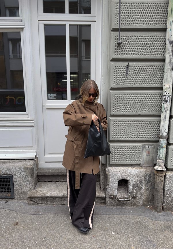 Woman wearing brown trench coat and black wide pants with white stripes, holding black handbag, standing on sidewalk near a building entrance.