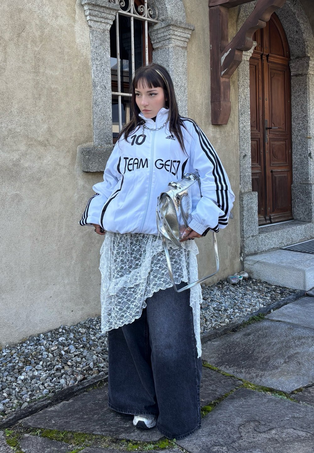 Biała kurtka sportowa z czarnymi paskami i napisem, warstwowy top koronkowy, ciemne szerokie dżinsy i srebrna torba. Tło ze ściany kamiennej.