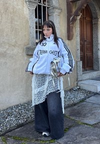 Biała kurtka sportowa z czarnymi paskami i napisem, warstwowy top koronkowy, ciemne szerokie dżinsy i srebrna torba. Tło ze ściany kamiennej.