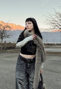 Zwart grafisch T-shirt met schedelontwerp, grijze distressed jeans met wijde pijpen, zilveren riem met studs en een nep bont sjaal over de schouder gedrapeerd.