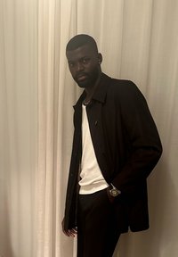 Άνδρας με σκούρο σακάκι, λευκό πουκάμισο και ρολόι στέκεται με το χέρι στην τσέπη μπροστά από φόντο με ανοιχτόχρωμη κουρτίνα.