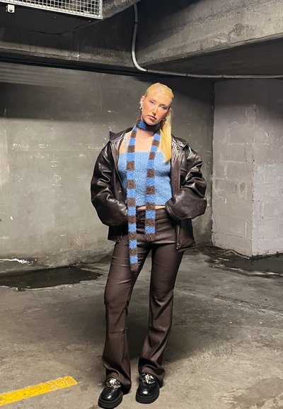 Chaqueta de cuero marrón oversize, top corta de felpa azul, bufanda de rayas, pantalones de rayas marrones y zapatos gruesos negros con acentos metálicos.