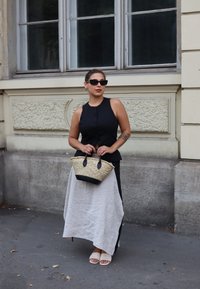 Femeie îmbrăcată cu un top negru fără mâneci, fustă asimetrică bej, sandale albe și ochelari de soare, ținând o geantă împletită pe trotuarul orașului.