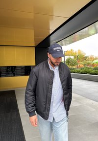 Jachetă bomber neagră peste o cămașă cu dungi alb-albastre, asortată cu blugi deschis la culoare și o șapcă închisă, stând pe o suprafață modernă în aer liber.