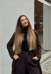 Jeune femme aux cheveux longs ombrés, portant un manteau foncé et un col roulé noir, debout en plein air, les mains dans les poches et souriant.