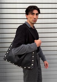 Sort læder skuldertaske med sølvfarvede kuppelnitter langs kanterne. Modellen har en sort top og grå ærmer på og fremviser en struktureret form.