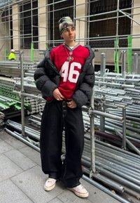 Jeune personne portant un bonnet camouflage, un maillot de sport rouge, une veste matelassée noire, un pantalon large noir et des baskets blanches, debout près d'un échafaudage métallique.