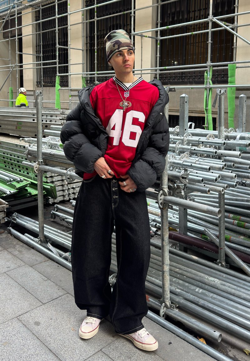 Jeune personne portant un bonnet camouflage, un maillot de sport rouge, une doudoune noire, un pantalon large noir et des baskets blanches, debout près d'un échafaudage métallique.
