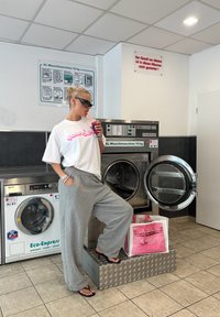 Λευκό oversize t-shirt με ροζ κείμενο, γκρίζες φόρμες, μαύρα γυαλιά ηλίου. Μηχανές πλυντηρίων στο φόντο, ροζ και άσπρη τσάντα σε μεταλλική πλατφόρμα.