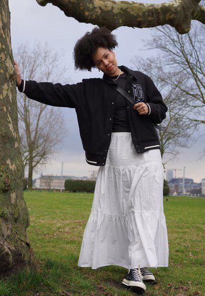 Chaqueta varsity negra con detalles en blanco, sobre una camiseta negra, combinada con una falda larga blanca de capas y zapatillas negras, posando frente a un árbol.