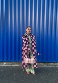 Femme debout devant un mur en tôle ondulée bleue, portant un manteau à carreaux rose et noir, une tenue verte à motifs, tenant un sac à main rose clair.