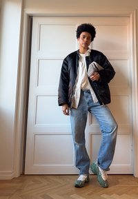 Μαύρο μπουφάν, λευκό πουκάμισο, ανοιχτόχρωμο τζιν και πράσινα-άσπρα αθλητικά παπούτσια. Κρατάει μια γκρι βελούδινη τσάντα. Ουδέτερο φόντο.