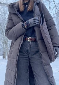 Personne portant un long manteau matelassé gris, un pull en maille noir, des gants noirs, un jean foncé avec une ceinture marron, et transportant un sac fourre-tout matelassé dans la neige.