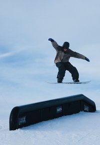 Snowboardeur en tenue noire en plein saut au-dessus d'un rail noir marqué des logos "LAAX" et "Swatch" sur une pente enneigée sous un ciel pâle.