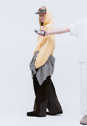 Prevelika rumena kapuca z kapuco. Rjave in sive hlače širšega kroja z karo srajco zavezano okoli pasu. Karirana kapica in prenosna naprava.