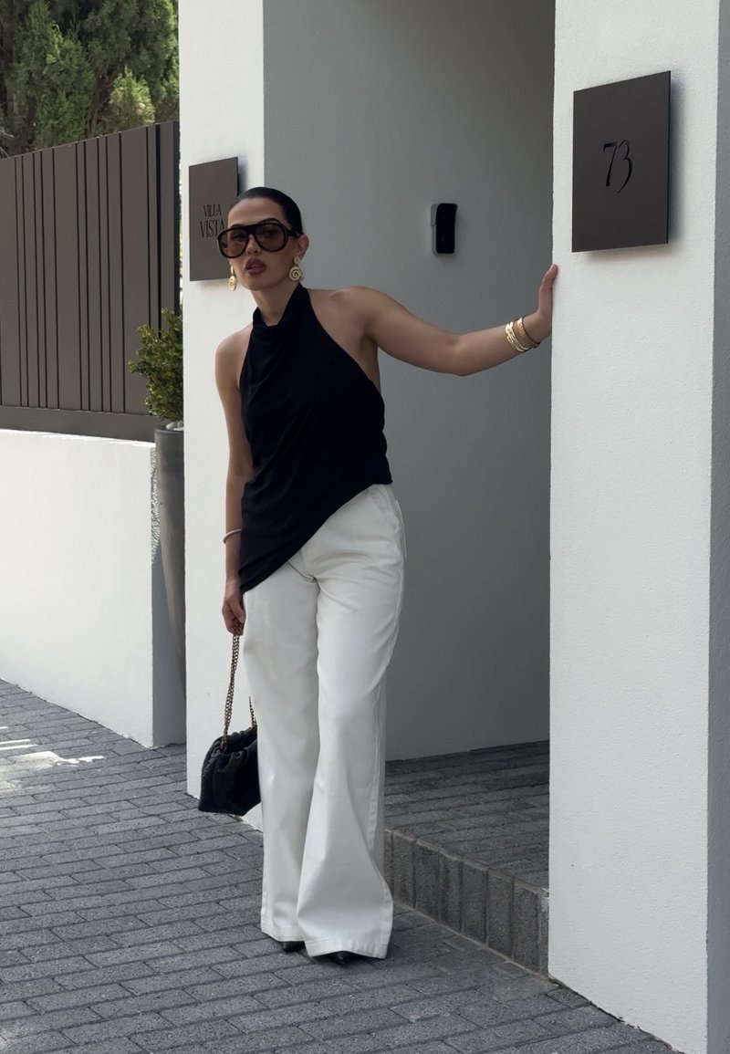 Femme portant un haut noir sans manches et un pantalon blanc, appuyée contre un mur blanc près d'une plaque noire avec le numéro 73, tenant un sac à main noir.