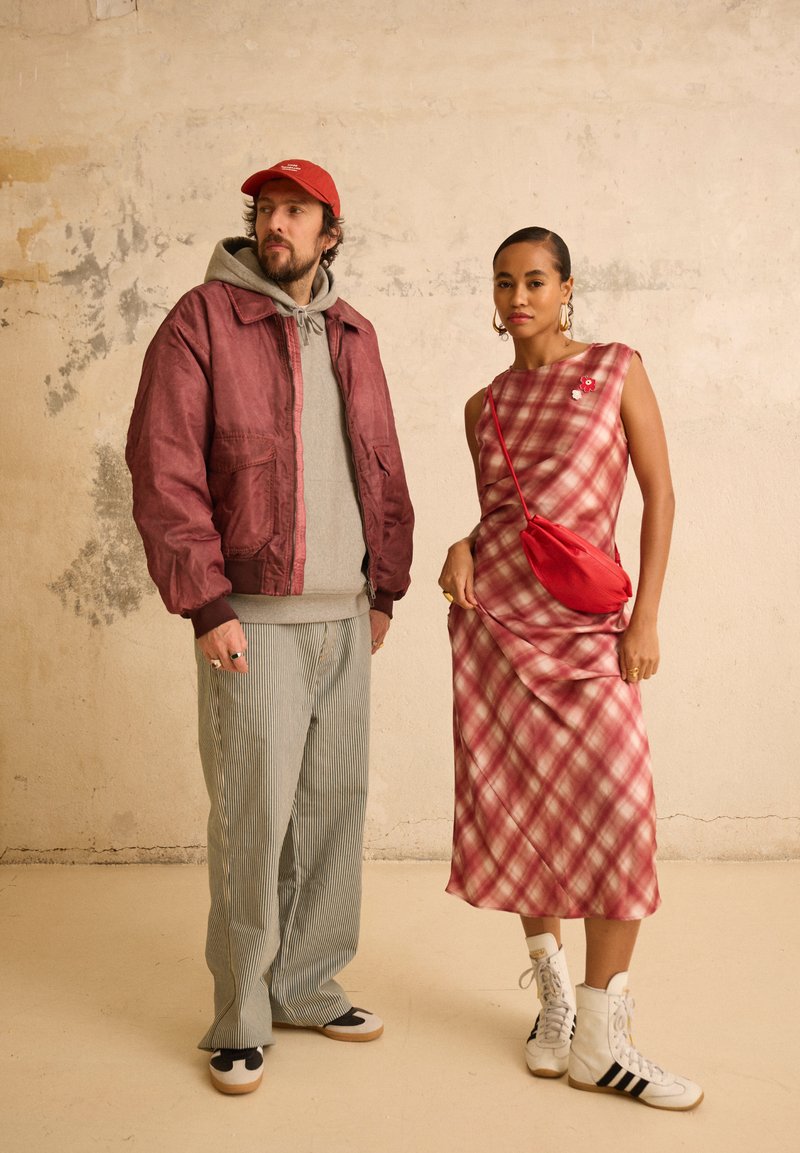 Bărbat cu jachetă roșie, hanorac gri, pantaloni cu dungi și șapcă stă lângă o femeie în rochie roșu în carouri și adidași înalți albi, cu geantă roșie purtată în diagonală.